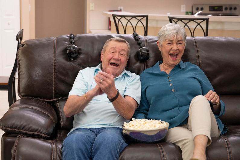 Joyful senior couple laughing on a couch with ChairSpeakers attached to their furniture, enjoying a shared audio experience.